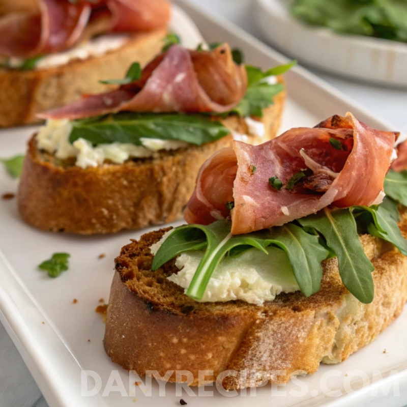 boursin-prosciutto-arugula-crostini (2) Appetizer crostini with creamy cheese, arugula, and prosciutto slices on toasted baguette.