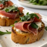 Crostini with whipped Boursin cheese, arugula, and folded prosciutto on a white tray.