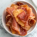Bowl of golden, crispy prosciutto slices stacked in a white dish on a striped napkin.