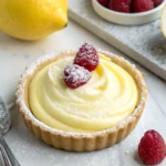 A tart shell filled with creamy lemon mousse, topped with raspberries and powdered sugar, set on a marble surface.