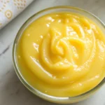 Glossy Meyer lemon curd swirled in a glass bowl on a marble surface.