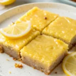 Four fudgy lemon blondies with a golden crust, topped with a lemon wedge and served on a speckled plate.