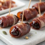 Baked prosciutto wrapped dates on a marble serving board, each pierced with a toothpick for easy serving.
