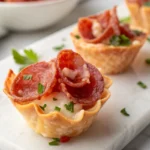 Salami cup appetizer bite filled with melted cheese and garnished with chopped parsley on a marble surface.